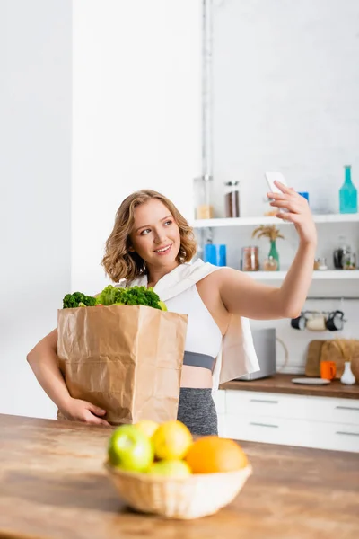 Enfoque selectivo de la mujer sosteniendo bolsa de papel con comestibles y tomando selfie - foto de stock