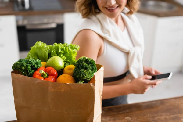 Selective focus of paper bag with groceries near woman holding smartphone with blank screen — Stock Photo