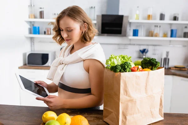 Junge Frau benutzt digitales Tablet mit leerem Bildschirm in der Nähe von Papiertüten mit Lebensmitteln — Stockfoto