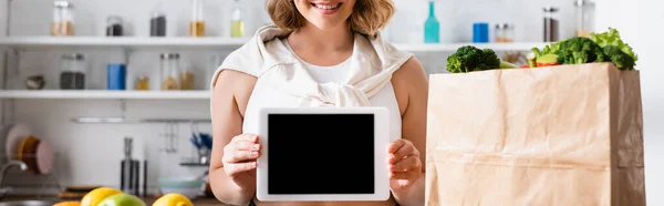 Панорамный урожай женщины с цифровым планшетом с чистым экраном рядом с бумажным пакетом с продуктами — стоковое фото