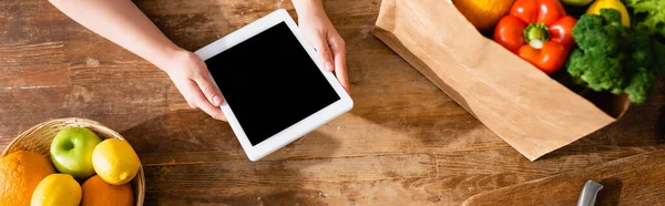 Concepto panorámico de mujer sosteniendo tableta digital con pantalla en blanco cerca de bolsa de papel con comestibles y frutas - foto de stock