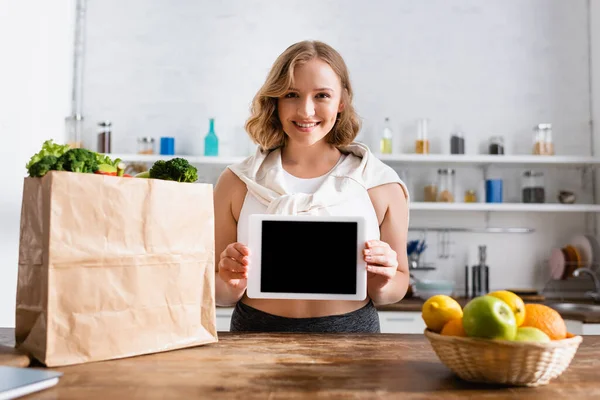 Foco seletivo da mulher segurando tablet digital com tela em branco perto de saco de papel com mantimentos e frutas — Fotografia de Stock