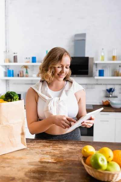 Foco seletivo da mulher usando tablet digital perto de saco de papel com mantimentos e frutas — Fotografia de Stock