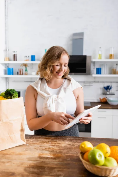 Messa a fuoco selettiva della donna utilizzando tablet digitale vicino sacchetto di carta con generi alimentari e frutta — Foto stock