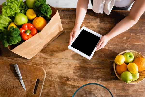 Vista superior de la mujer sosteniendo tableta digital con pantalla en blanco cerca de la bolsa de papel con comestibles y frutas - foto de stock