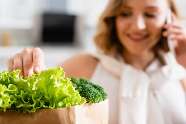 Selective focus of woman touching fresh lettuce near broccoli while talking on smartphone — Stock Photo