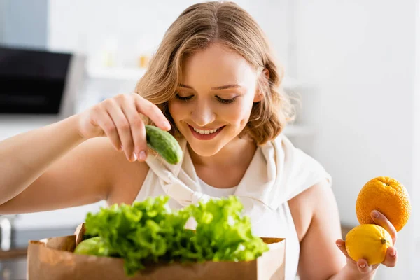 Selective focus of woman holding cucumber, lemon and orange while looking at paper bag with lettuce — Stock Photo