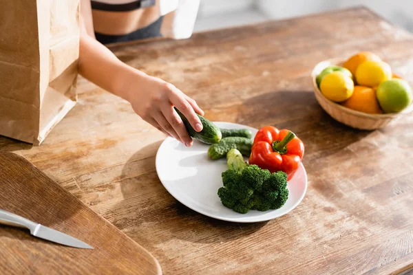 Partial view of young woman holding cucumber near plate with broccoli and red bell pepper — Stock Photo