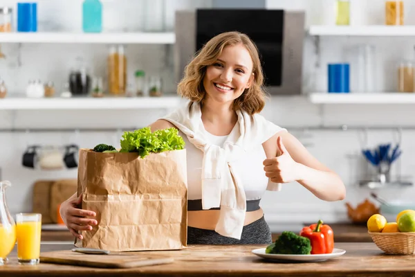 Junge Frau zeigt Daumen hoch neben Papiertüte mit Salat und Schale mit Früchten — Stockfoto