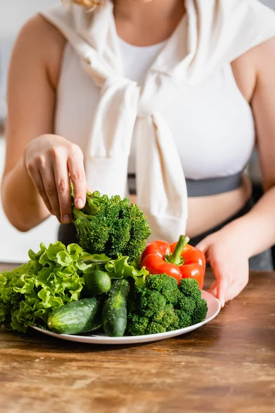 Vista recortada de la mujer sosteniendo brócoli fresco cerca de verduras maduras en el plato - foto de stock