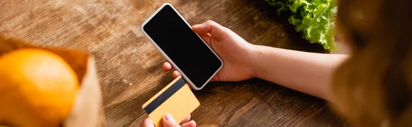 Cultivo panorámico de mujer sosteniendo teléfono inteligente con pantalla en blanco y tarjeta de crédito cerca de lechuga y naranja - foto de stock