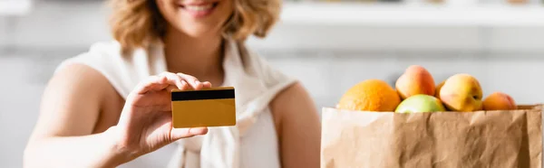 Plano panorámico de mujer sosteniendo tarjeta de crédito cerca de bolsa de papel con comestibles - foto de stock