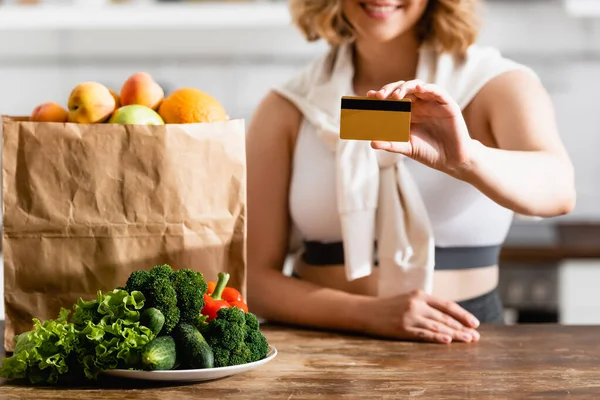 Vista parcial de la mujer que sostiene la tarjeta de crédito cerca de la bolsa de papel y el plato con verduras en la mesa - foto de stock