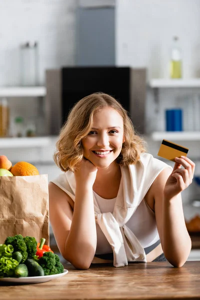 Жінка тримає кредитну картку біля паперового мішка і тарілки з овочами на столі — стокове фото