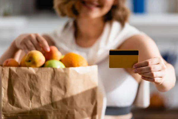 Cropped view of woman holding credit card near paper bag with fruits — Stock Photo