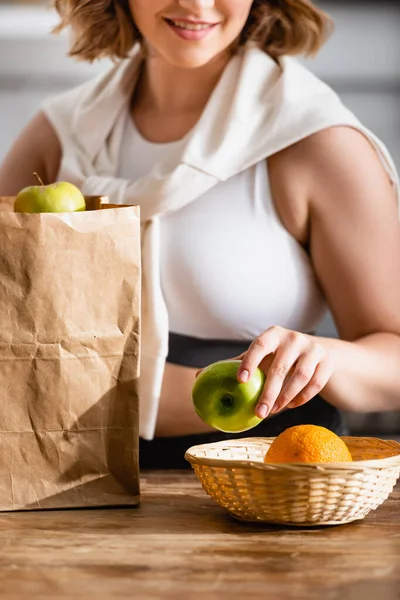 Partial view of woman holding apple near paper bag and fruits — Stock Photo