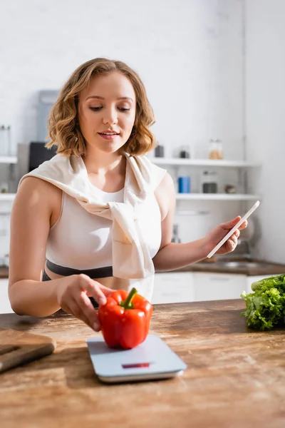 Mujer tocando pimiento en balanzas de cocina y sosteniendo teléfono inteligente - foto de stock