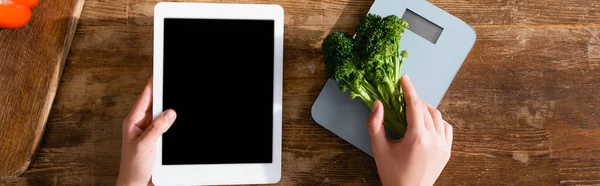 Horizontal image of woman holding broccoli near kitchen scales and digital tablet with black screen — Stock Photo