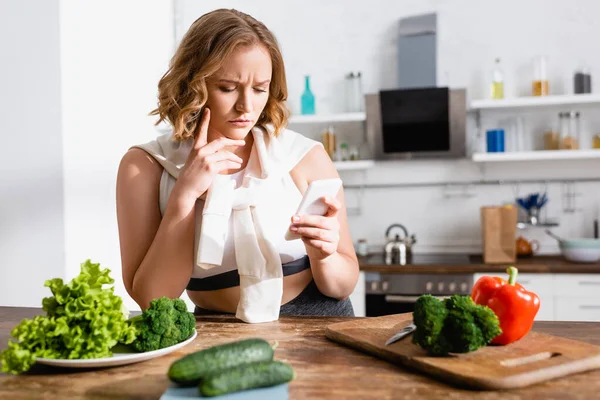 Selective focus of upset woman using smartphone near vegetables in kitchen — Stock Photo