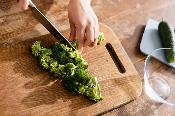 Visão parcial da mulher cortando brócolis fresco na tábua de corte perto de balanças de cozinha com pepinos — Fotografia de Stock
