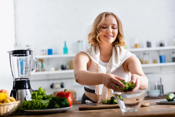 Selective focus of young woman putting broccoli in bowl near vegetables — Stock Photo