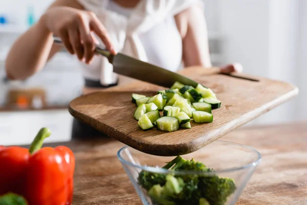 Vista recortada de la mujer sosteniendo tabla de cortar con pepino en rodajas cerca del tazón con brócoli - foto de stock