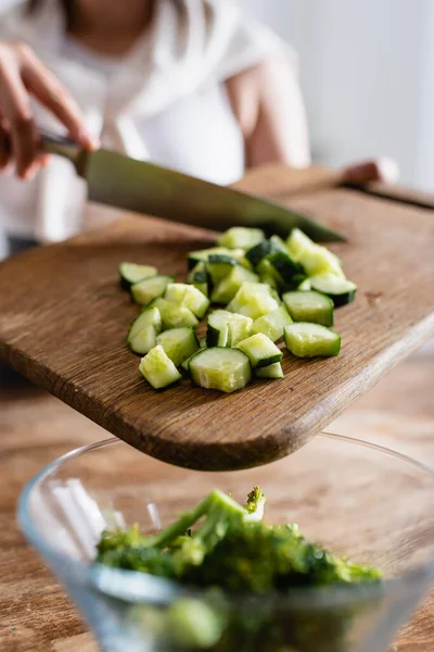 Vista cortada da mulher segurando placa de corte com pepino fatiado perto da tigela com brócolis fresco — Fotografia de Stock