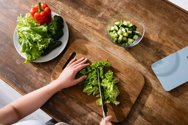 Von oben: Frau schneidet grünen Salat auf Schneidebrett in der Nähe von Küchenwaagen und ganzen Gemüsesorten — Stockfoto