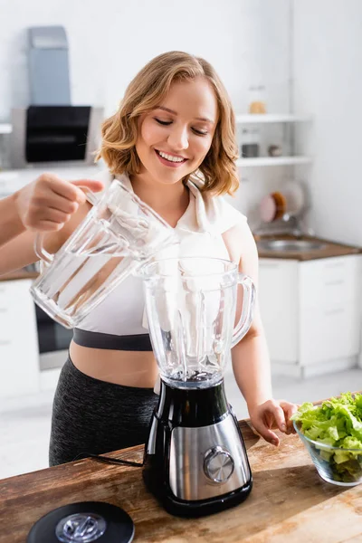 Junge Frau gießt Wasser in Mixer neben Schüssel mit Salat — Stockfoto