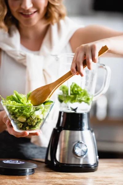 Vista ritagliata della donna che tiene ciotola con lattuga fresca e cucchiaio di legno vicino frullatore — Foto stock