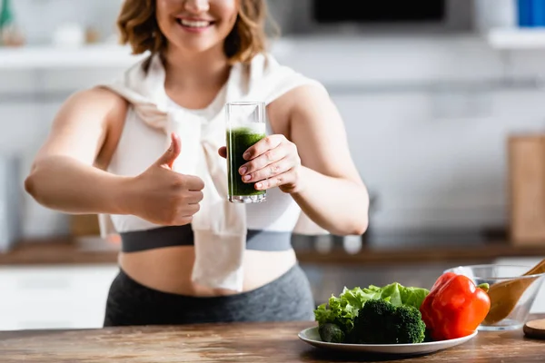 Vista recortada de la joven mujer sosteniendo un vaso de batido verde mientras muestra el pulgar hacia arriba - foto de stock