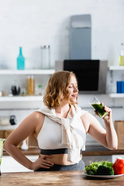 Jeune femme tenant un verre de smoothie vert tout en se tenant avec la main sur la hanche — Photo de stock