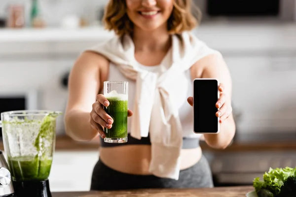 Vista recortada de mujer joven sosteniendo vidrio de batido verde y teléfono inteligente con pantalla en blanco - foto de stock