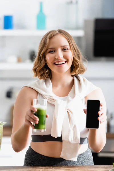 Enfoque selectivo de la mujer joven sosteniendo vidrio de batido verde y teléfono inteligente con pantalla en blanco - foto de stock