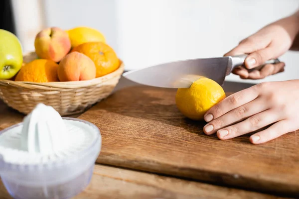 Vue recadrée d'une femme coupant du citron sur une planche à découper près de fruits et de presse-fruits — Photo de stock