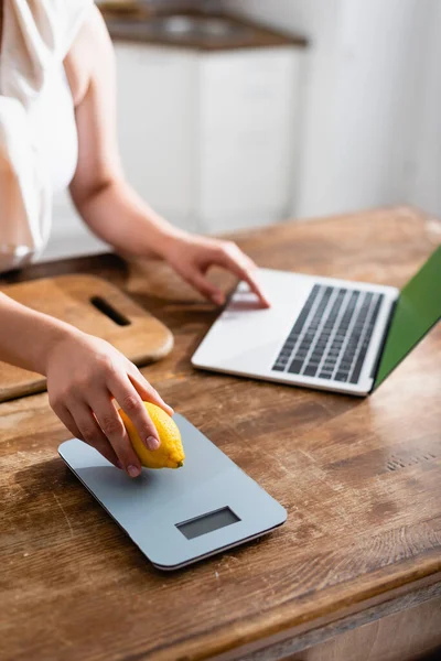 Cropped view of woman holding lemon near kitchen scales and using laptop — Stock Photo