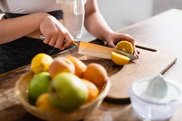 Vista recortada de la joven mujer cortando limón fresco en la tabla de cortar cerca de frutas y exprimidor - foto de stock