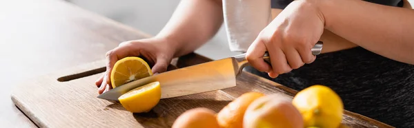 Panoramic crop of young woman cutting fresh lemon on chopping board near fruits and juicer — Stock Photo