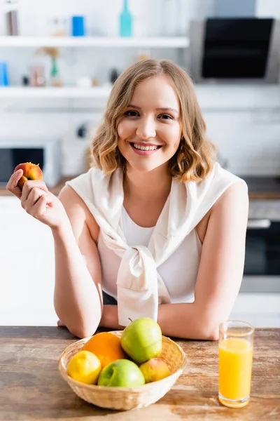 Femme tenant pêche fraîche tout en regardant la caméra dans la cuisine — Photo de stock