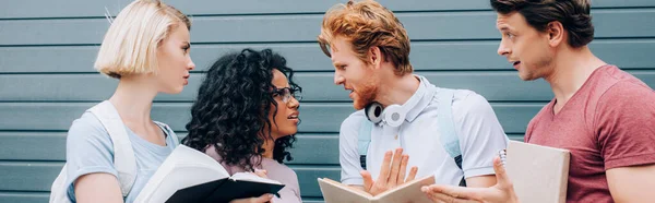 Panoramic concept of serious multicultural students talking while standing with open books on street — Stock Photo