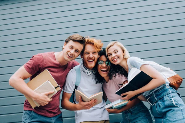 Estudiantes multiculturales emocionados mirando a la cámara mientras sostienen libros de texto en la calle - foto de stock