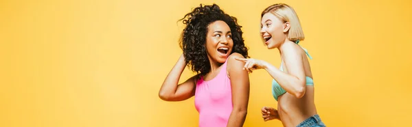 Horizontales Bild einer aufgeregten blonden Frau, die mit dem Finger in die Nähe eines afrikanisch-amerikanischen Freundes zeigt, der Haare isoliert auf gelb berührt — Stockfoto