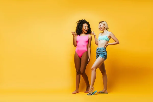 Full length view of young african american woman and blonde friend with hand on hip pointing with fingers on yellow — Stock Photo