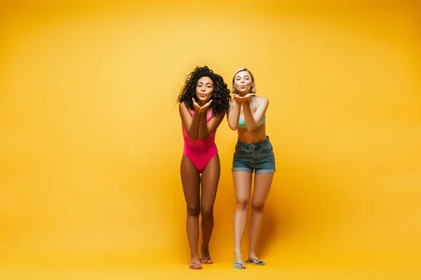 Full length view of multicultural women blowing air kisses at camera on yellow — Stock Photo