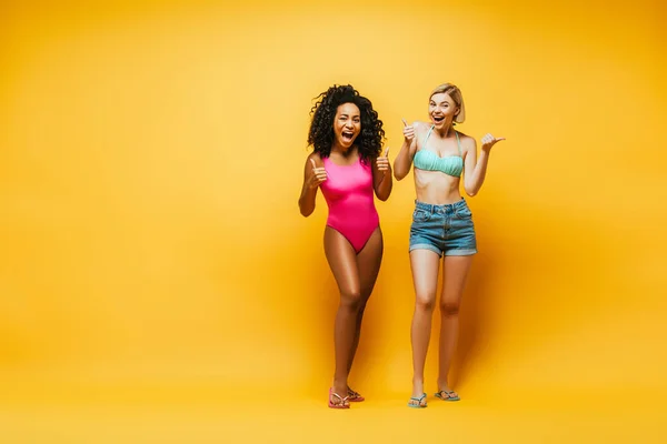 Full length view of excited interracial women in summer outfit showing thumbs up while looking at câmera on yellow — Fotografia de Stock