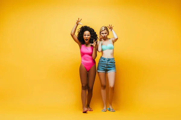 Молоді міжрасові жінки в літньому вбранні показують жести, дивлячись на камеру на жовтому — стокове фото