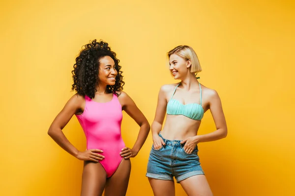 Curly african american woman with hands on hips near blonde friend with hands in pockets on yellow — Stock Photo