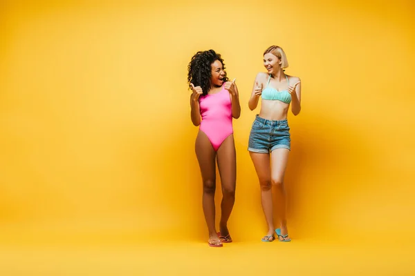 Vista completa de excitado interracial las mujeres mostrando los pulgares hacia arriba y mirando el uno al otro en amarillo - foto de stock