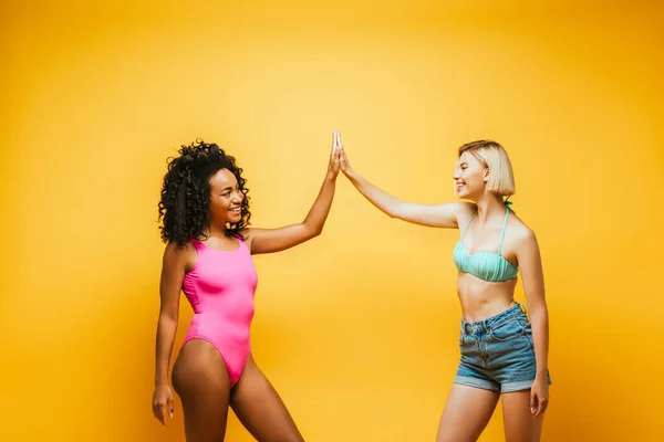 Junge afrikanisch-amerikanische Frau im Badeanzug gibt High Five an blonde Freundin in Shorts auf gelb — Stockfoto
