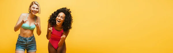 Immagine orizzontale di eccitate donne interrazziale ridendo e indicando con le dita la fotocamera isolata sul giallo — Foto stock